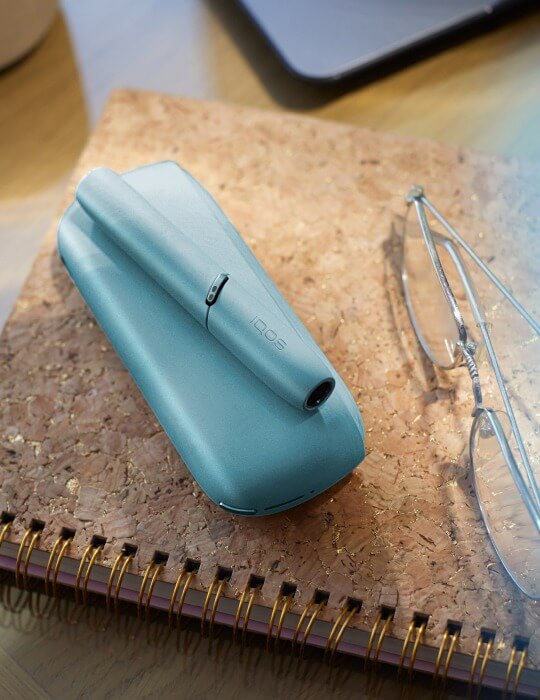 Turquoise IQOS Originals Duo device on a notebook next to glasses.