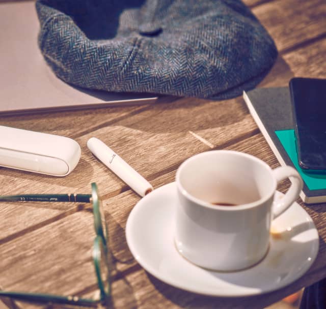 IQOS 3 DUO on a table next to a cup of coffee and a cap