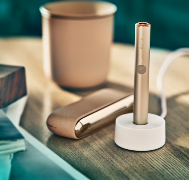 Close up of gold khaki IQOS ILUMA device charging, next to books and a cup.