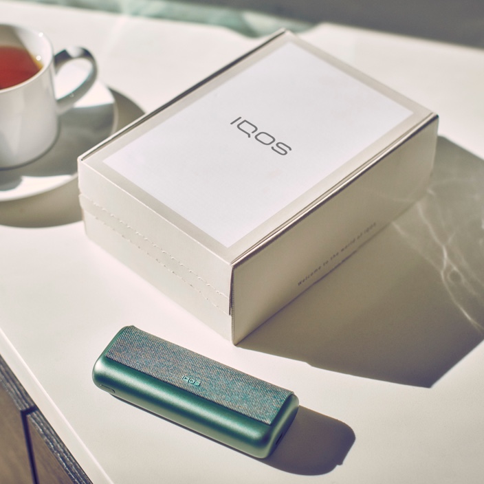 A jade green IQOS ILUMA PRIME Pocket Charger and an IQOS box on a table.