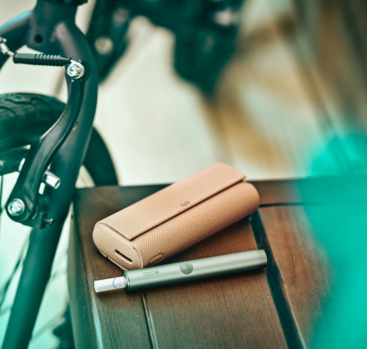 מחזיק IQOS ILUMA PRIME בצבע 
ירוק ומטען נייד על שולחן ליד אופניים.