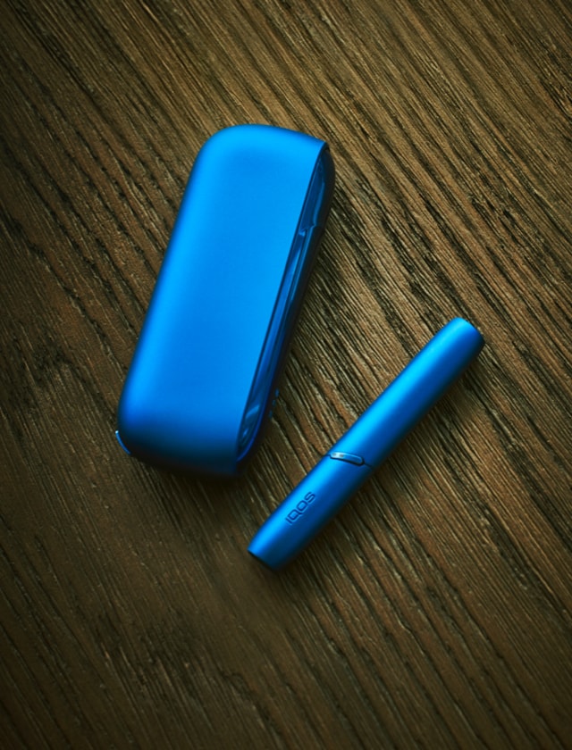 A blue IQOS device and holder on a wooden surface.
