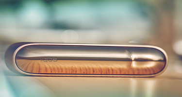 Two people at a table bar, one holds his IQOS device and the other uses his phone